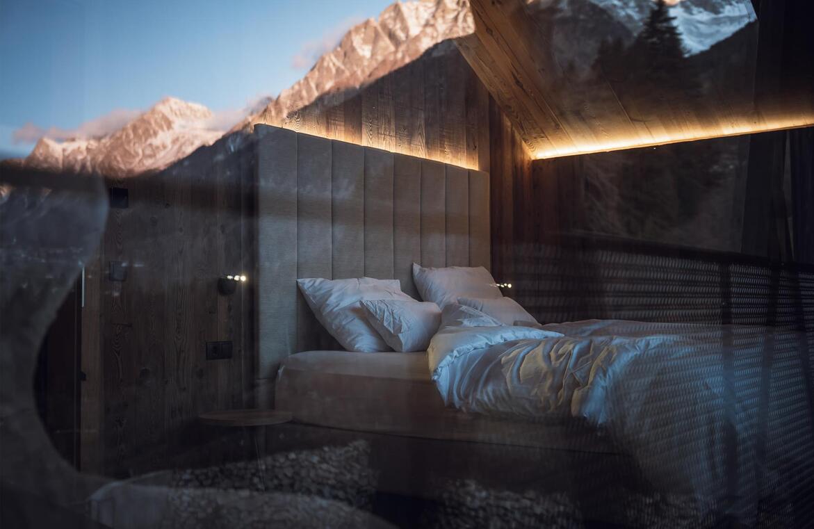 Chalet-Schlafzimmer mit Doppelbett, gepolstertem Kopfteil, indirekter Beleuchtung und Spiegelung der Berge in der Glasfront.