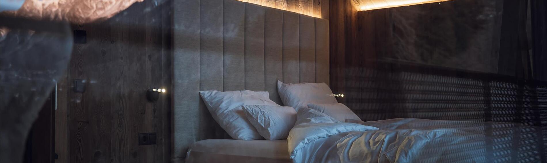 Chalet bedroom with a double bed, upholstered headboard, indirect lighting and mountains reflected in the glass.