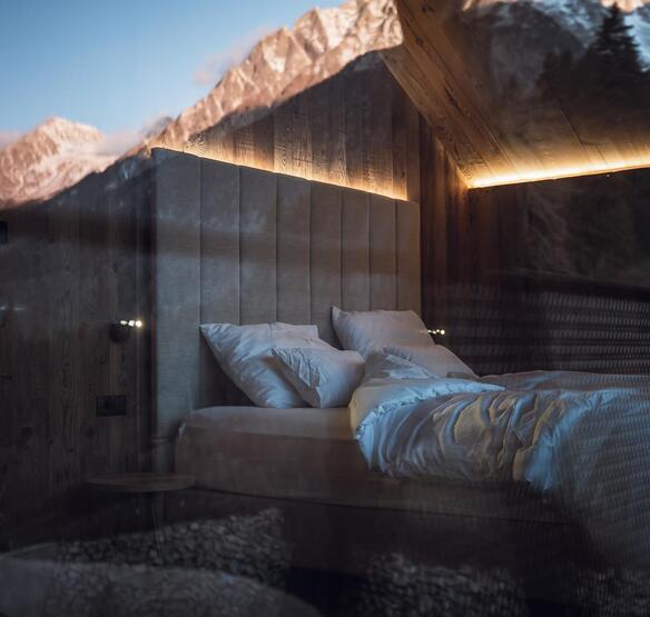 Camera da letto dello chalet con letto matrimoniale, testiera imbottita, illuminazione indiretta e montagne riflesse nella vetrata.